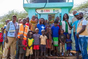 A maasai community and local action against hunger volunteers standing infront of a water station funded by UBS and action against hunger