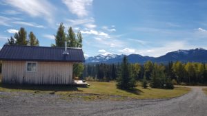 A small shack in a wide clearing of trees and mountains in the background