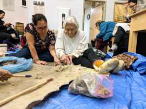 Two women sitting on the ground on top of a blue tarp learning how to dehair a bearded seal skin