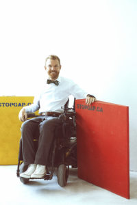 Full body shot of Luke Anderson, Founder and Executive Director of StopGap Foundation in his wheelchair in front of accessibility ramps with the STOPGAP.CA logo