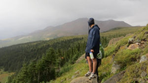 A man standing on the edge of a cliff over looking vast green forests and mountains in the background