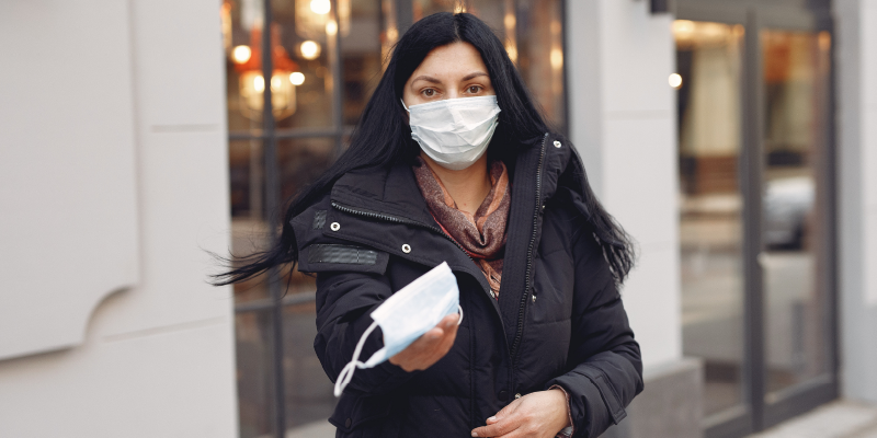 Woman wearing a mask with an outstretched hand holding out a mask to the camera man.